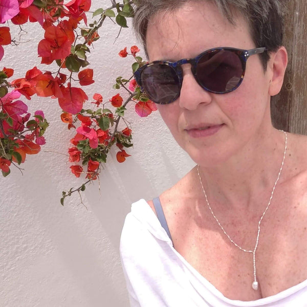 A minimal necklace made of fresh-water fragrant pearls, in the shape of a tie. A bigger pearl is at the lower end. Here photographed on our model in the streets of Paroikia.